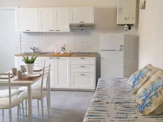 A compact kitchen and dining area with white cabinets, a stainless steel sink, a stove, and a small refrigerator. A table with chairs and a sofa with patterned cushions are also visible.