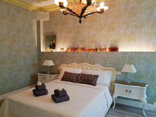 An elegantly decorated bedroom with a plush white bed, ornate bedside tables, lamps, and a classic textured wallpaper.