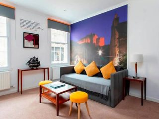 A modern apartment with a large photographic feature wall of Edinburgh Castle at dusk, complemented by a grey sofa with bright yellow cushions