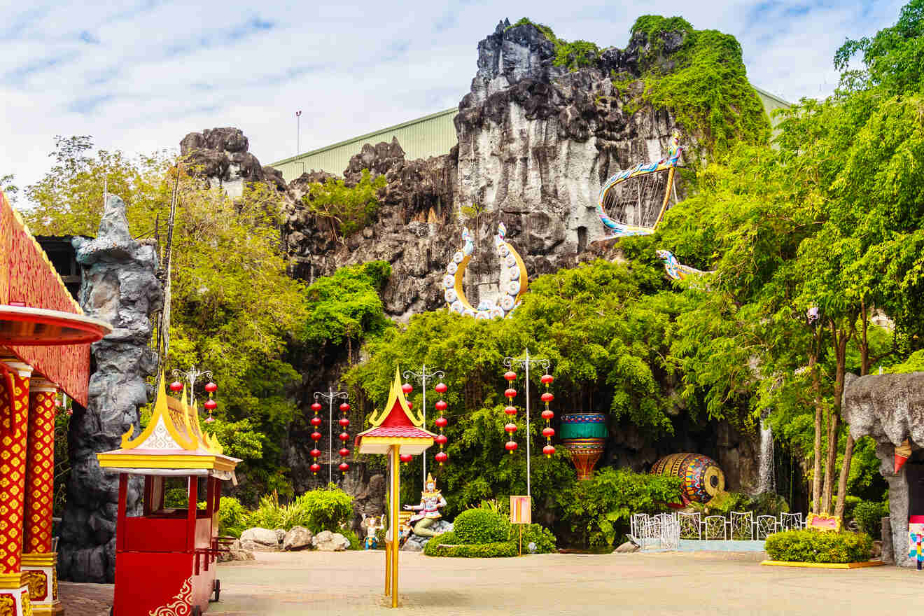 Traditional Thai cultural theme park with ornate red and gold structures, a large mountainous backdrop, and vibrant decorations, inviting visitors into a world of historic and mythological wonders.

