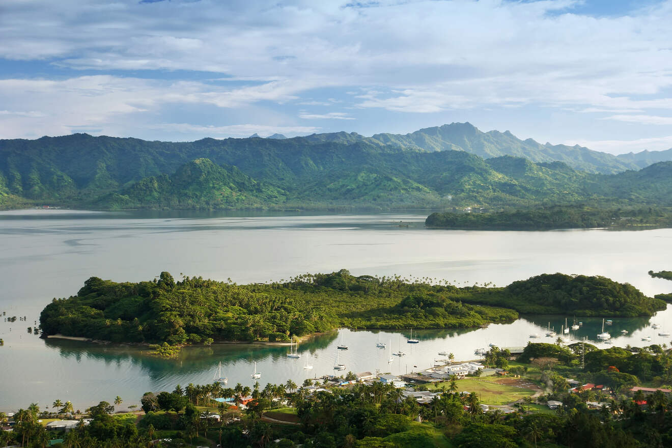A scenic panoramic view of a calm bay with a lush green island in the center, surrounded by mountains and moored sailboats, showcasing a serene and picturesque harbor