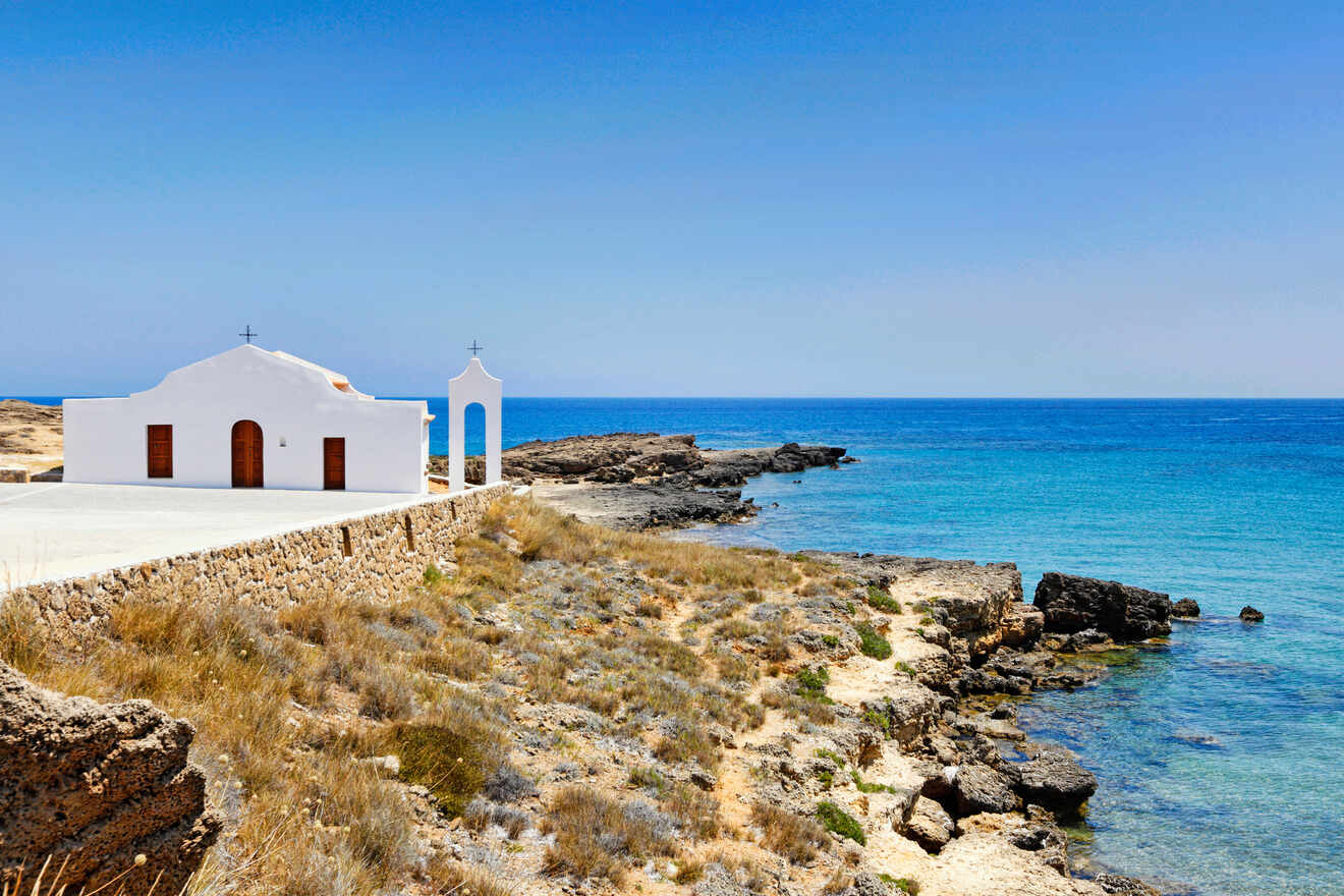 A small white chapel with an arched entrance overlooks a rocky coastline and clear blue sea under a bright blue sky.