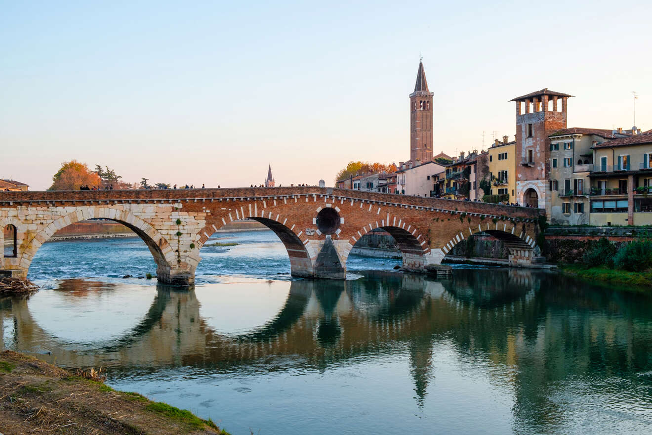 The historic Ponte Pietra bridge in Verona, Italy, crossing the tranquil Adige River during sunset, with warm lighting and clear skies