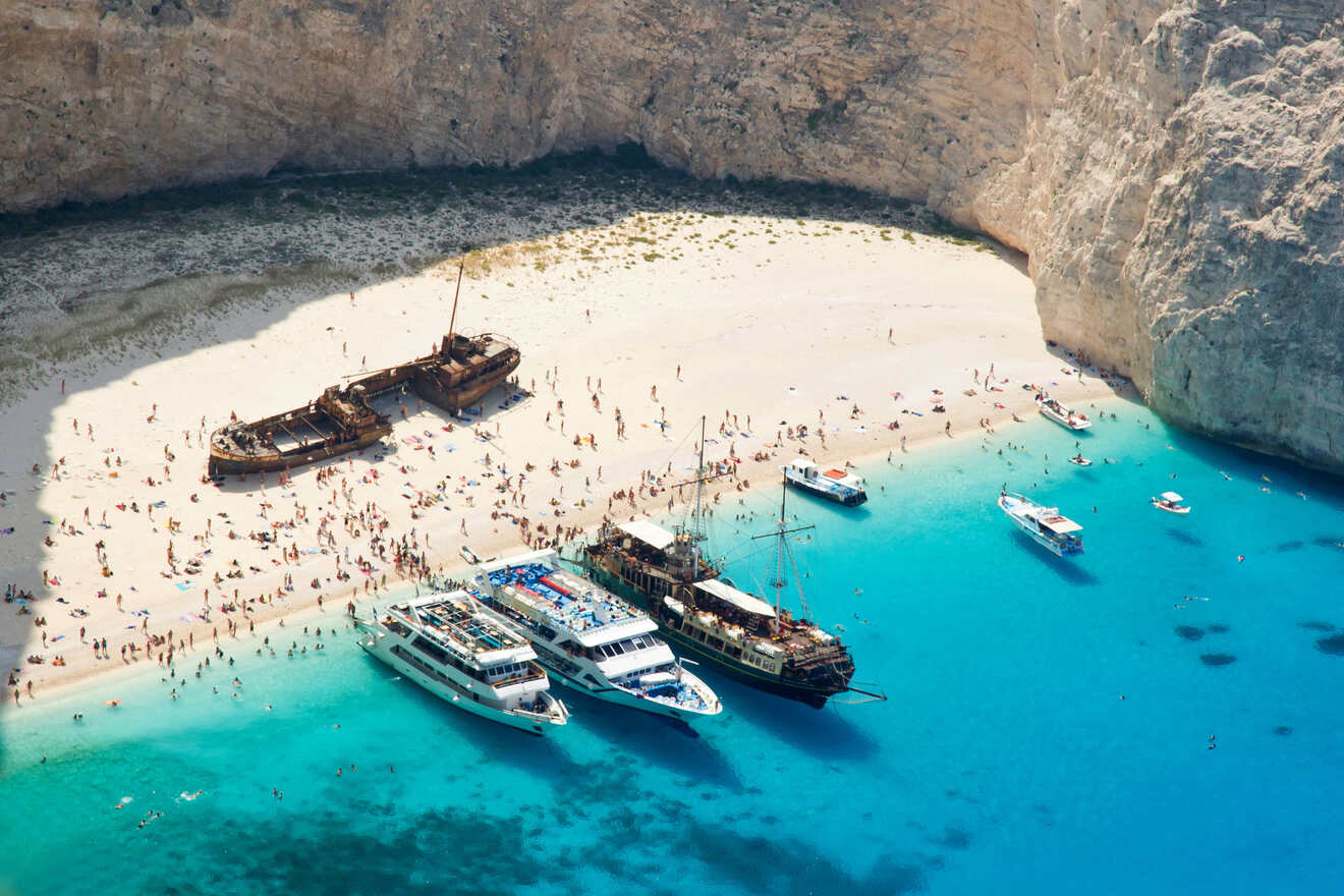 Aerial view of a beach with clear turquoise water, several boats docked, and people scattered along the sandy shore near rusty shipwrecks. Rocky cliffs surround the area.