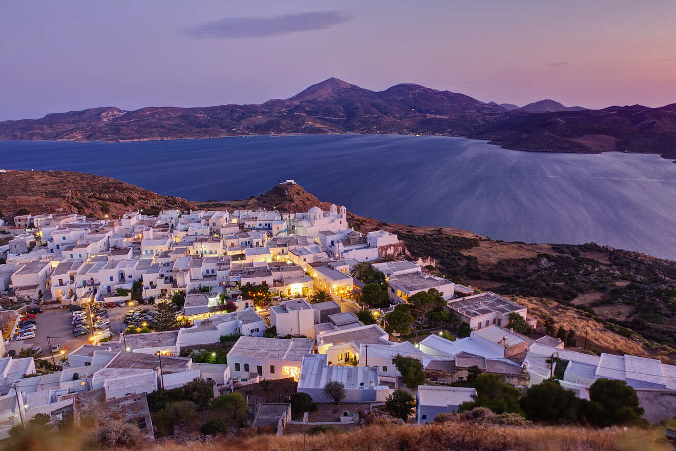 A stunning view of the Greek village Milos at dusk, with lights illuminating the white buildings and the calm sea in the background