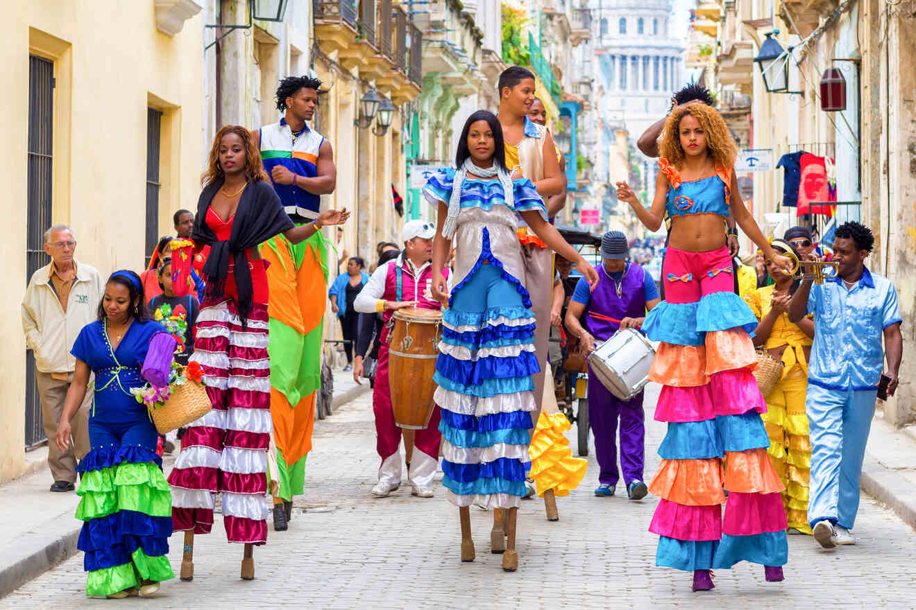 A group of people, some on stilts and others on foot, parade down a colorful street. They wear vibrant, multicolored costumes and are accompanied by musicians playing drums and other instruments.