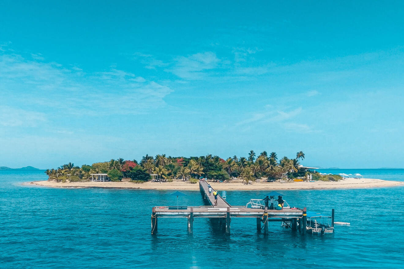 A distant view of a small, tranquil island with a dock extending into the clear blue waters, surrounded by lush greenery and a few scattered clouds in the sky