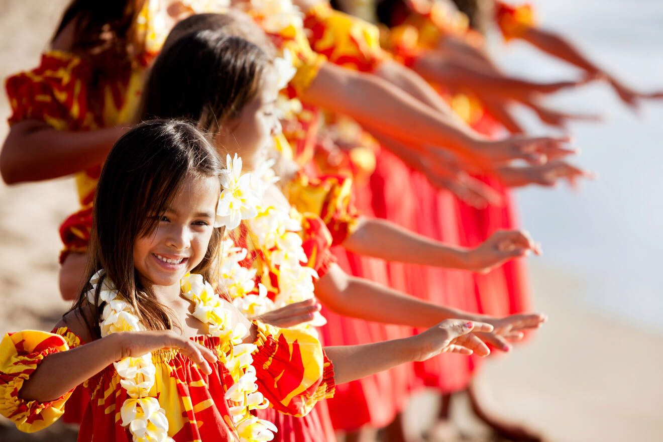 Girls in red and yellow dresses and floral necklaces perform a hula dance on a sandy beach.