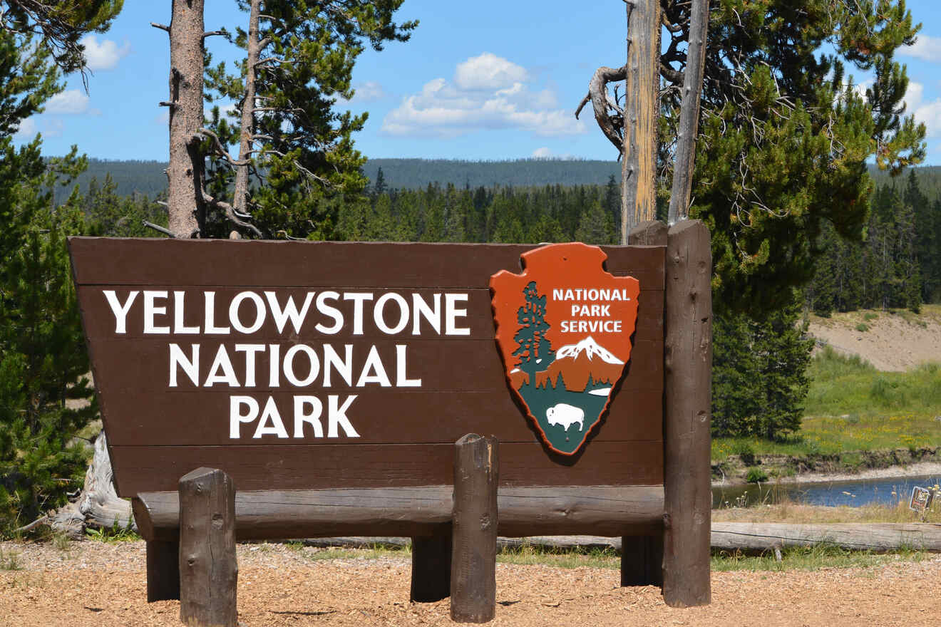 Sign reading "Yellowstone National Park" with National Park Service logo in a forested area under a blue sky.