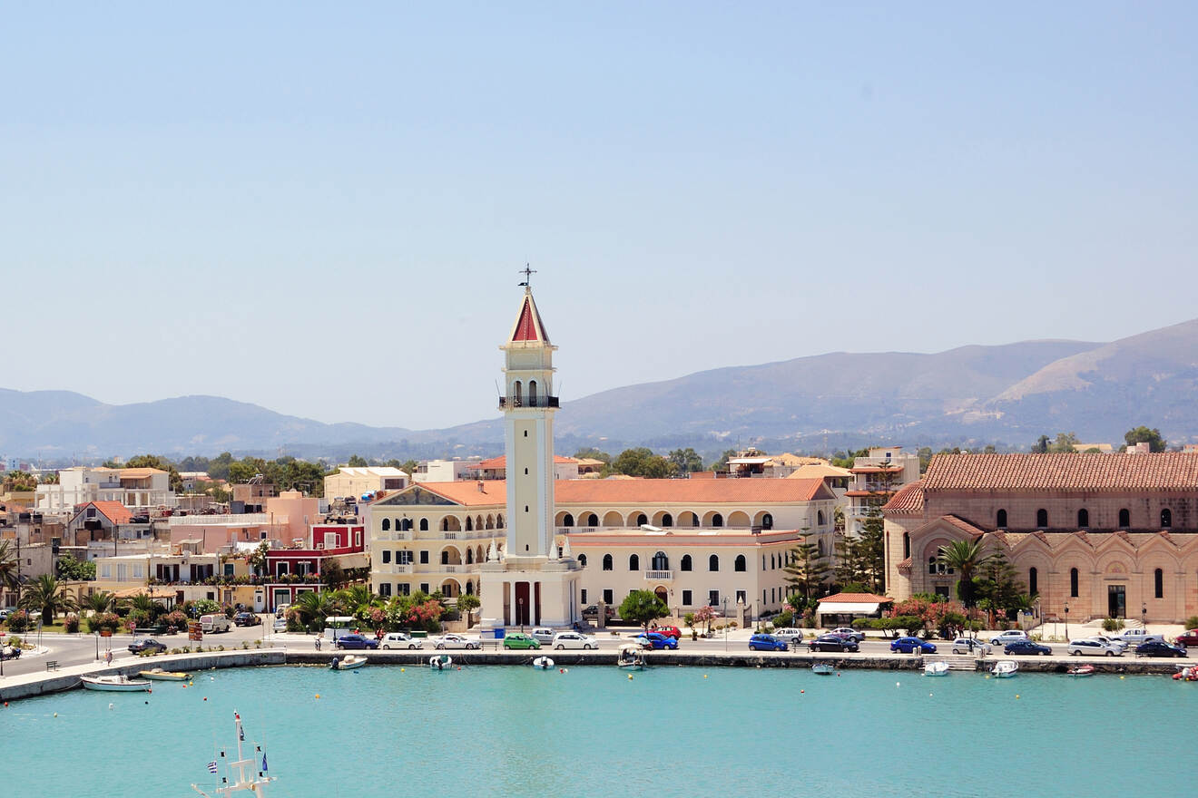 Coastal town with a prominent bell tower near the waterfront. Mountains in the background and boats lining the harbor.