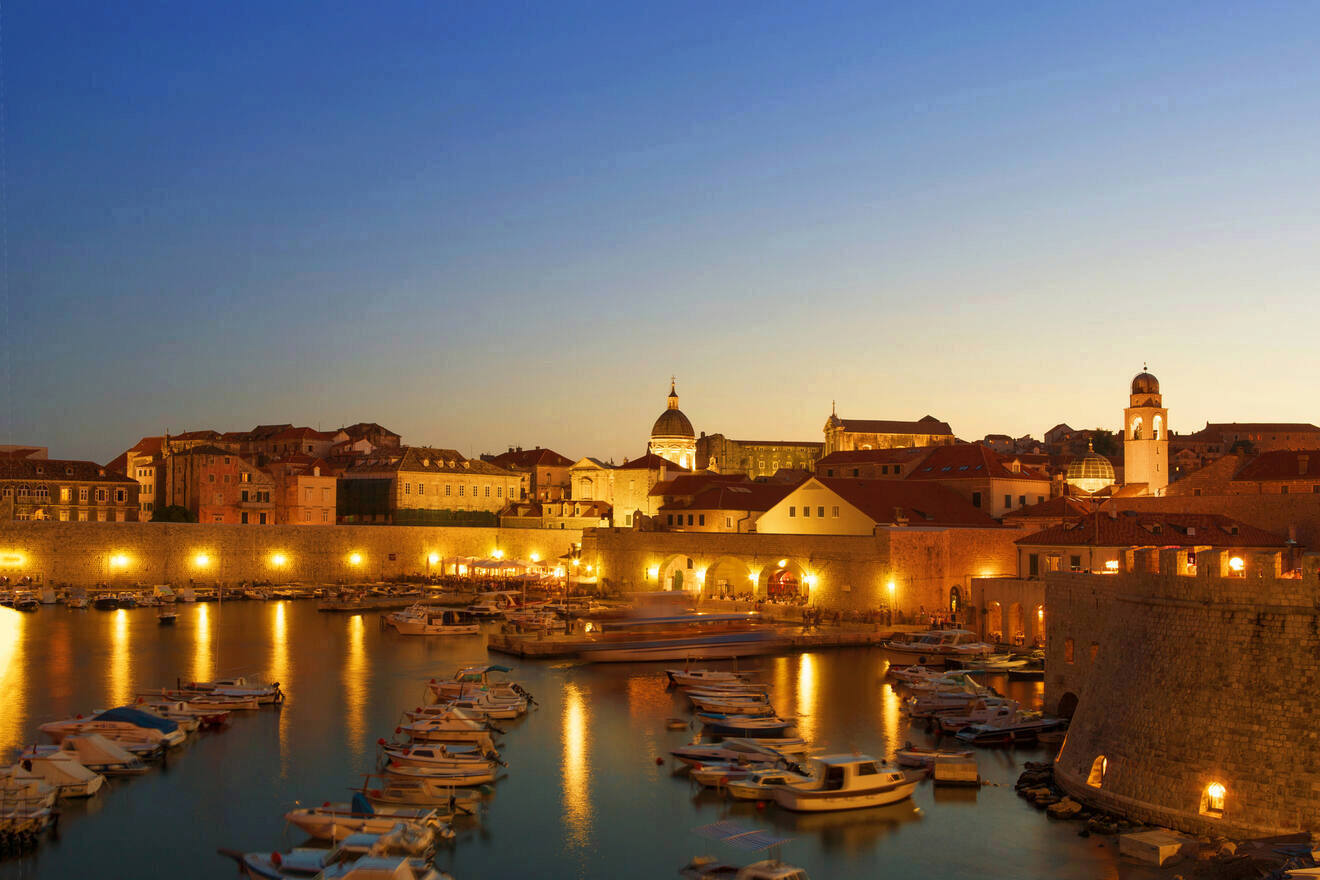 Dubrovnik's old town and harbor illuminated at dusk, with boats moored in calm waters and historic buildings lit up