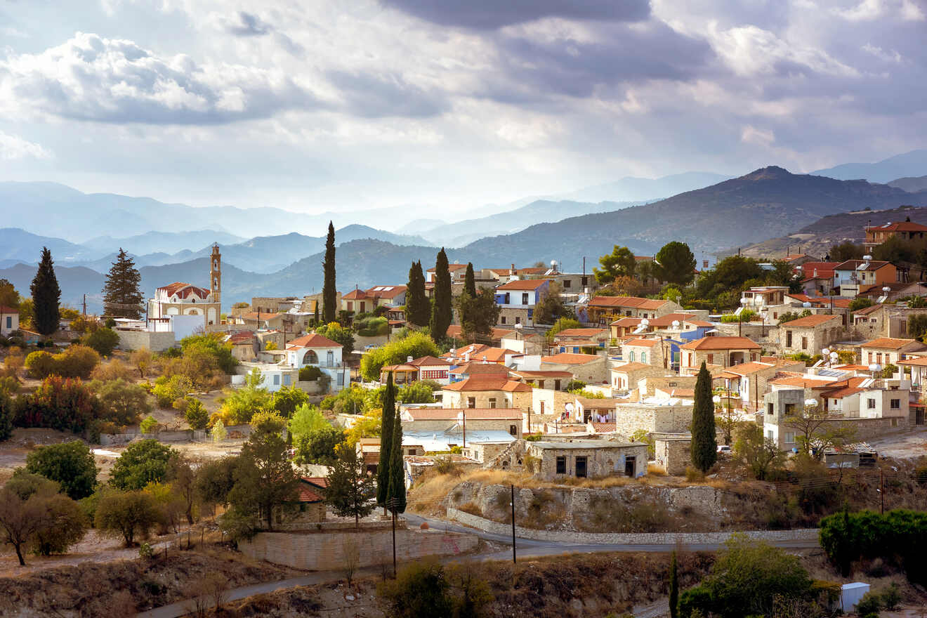 Scenic view of a traditional hillside village in Cyprus with a church, nestled among mountains under a cloudy sky, exuding rustic charm