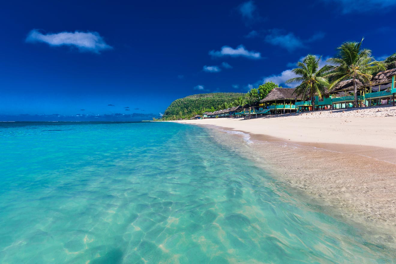 A pristine beach with turquoise waters along a lush tropical coastline, with thatched-roof structures under a clear blue sky