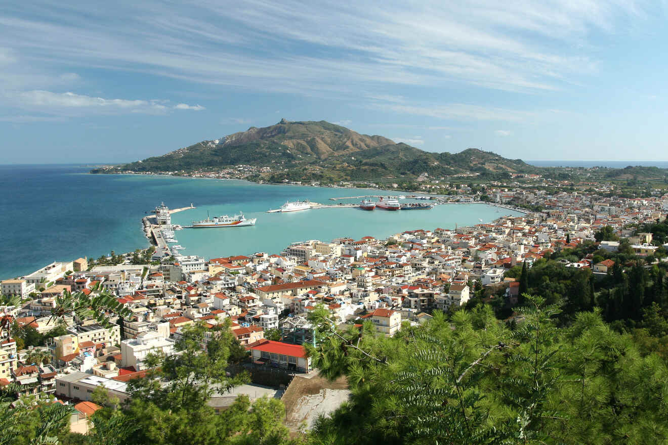 Coastal city view with a harbor, turquoise waters, several ships, densely packed buildings, and a mountainous backdrop under a blue sky.