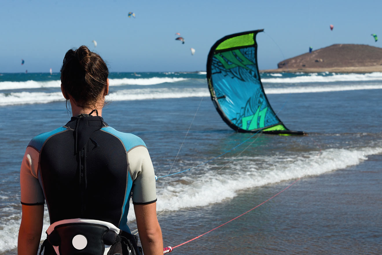 Woman in wetsuit and harness preparing for kitesurfing, gazing out at the sea with a blue and green kite in the water.