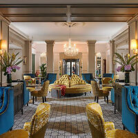 Elegant hotel lobby with blue and yellow armchairs, patterned floor, and a central yellow sofa under a grand chandelier