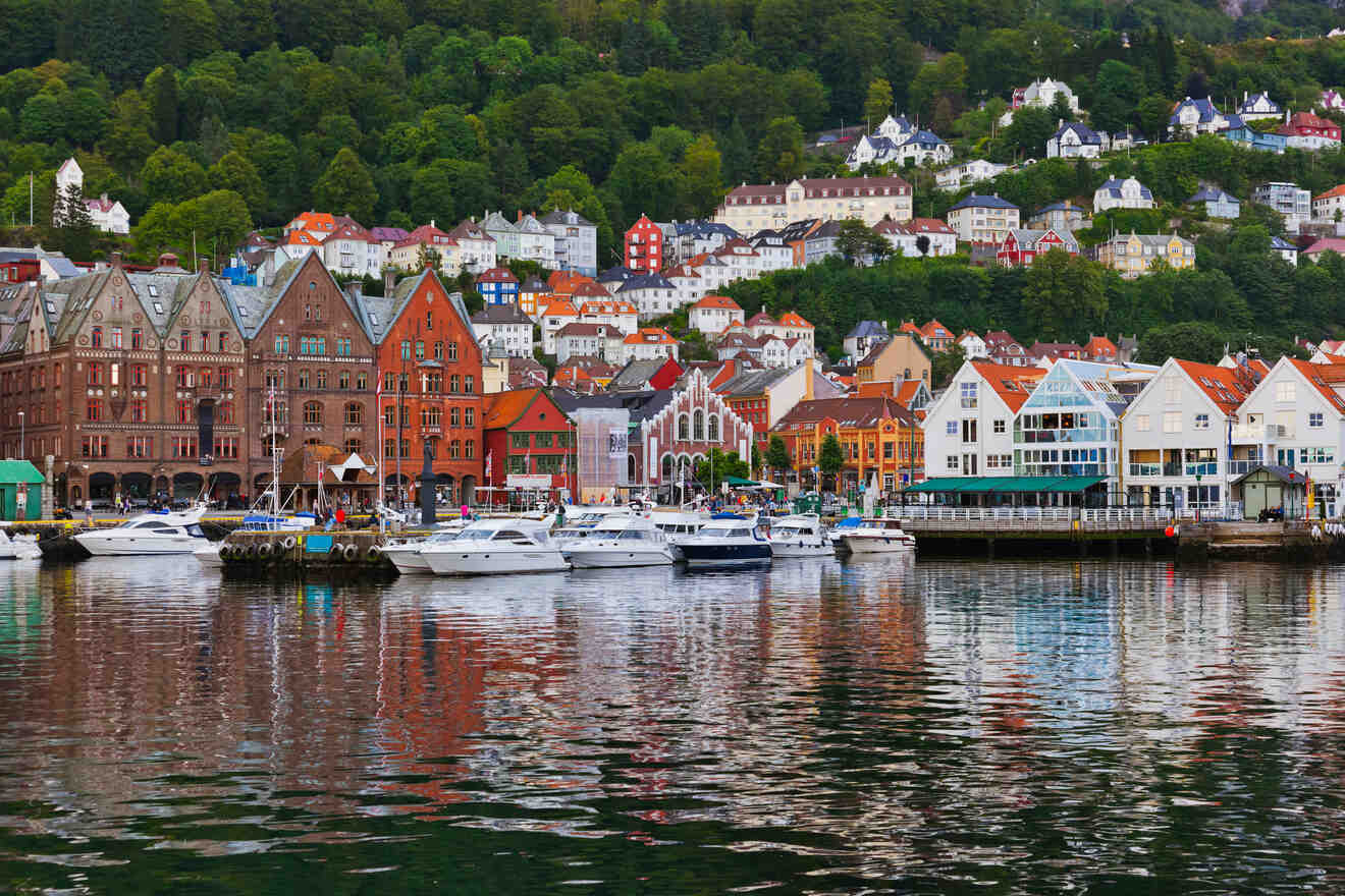 Colorful historical buildings line the waterfront of Bergen, Norway, with lush green hills in the background and luxury yachts moored in the harbor, reflecting a vibrant city life by the sea