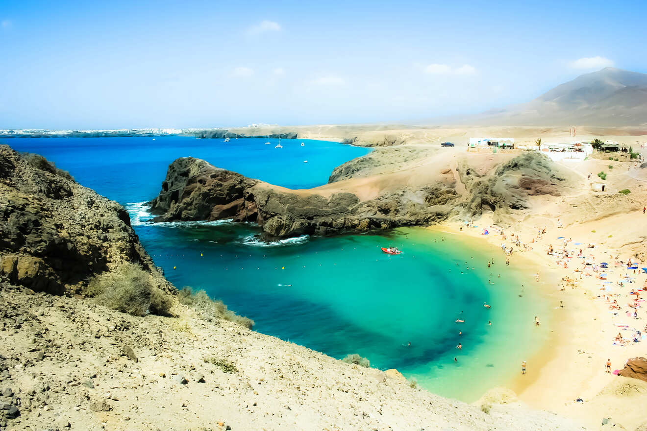 A picturesque cove on Lanzarote, Canary Islands, with turquoise waters and sandy beach surrounded by volcanic rocks, bustling with beachgoers and boats.