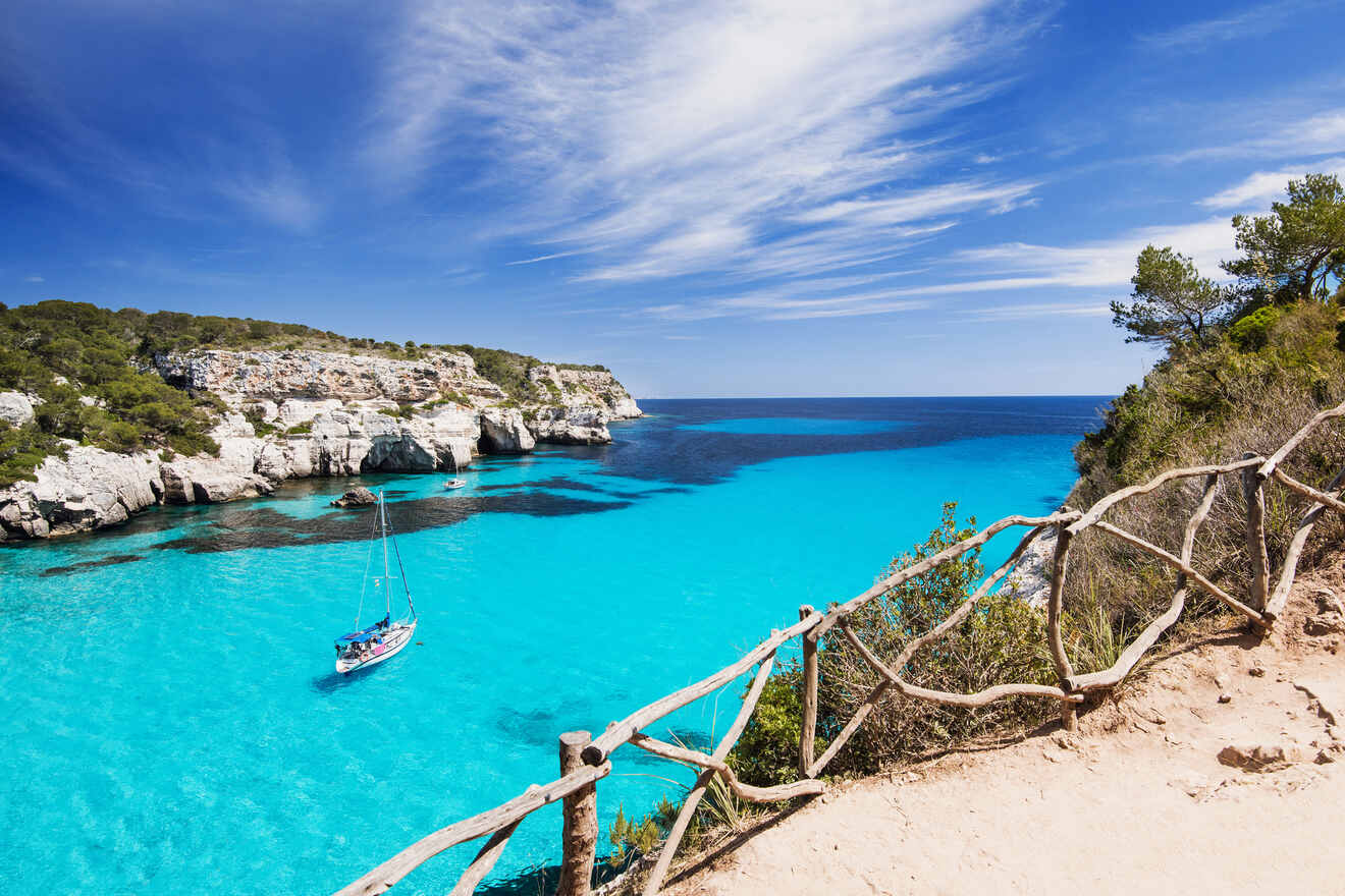 Overlooking Cala Macarella, Menorca, the view captures a wooden railing leading towards the stunning cove with turquoise waters and moored sailboats, encapsulating the island’s allure