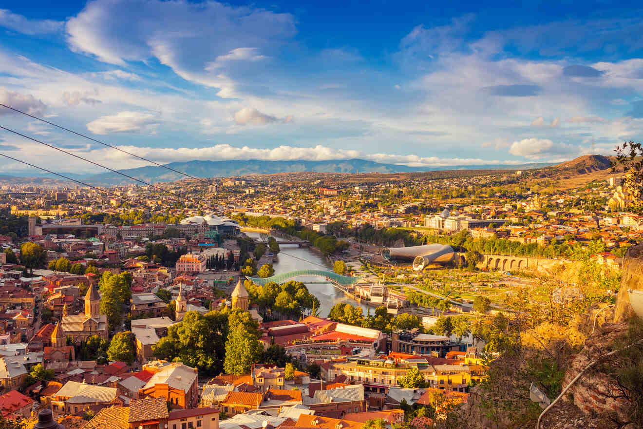 A panoramic view of Tbilisi with the Peace Bridge and Rike Park visible, showcasing the city's blend of historical architecture and modern landmarks under a golden sunset.