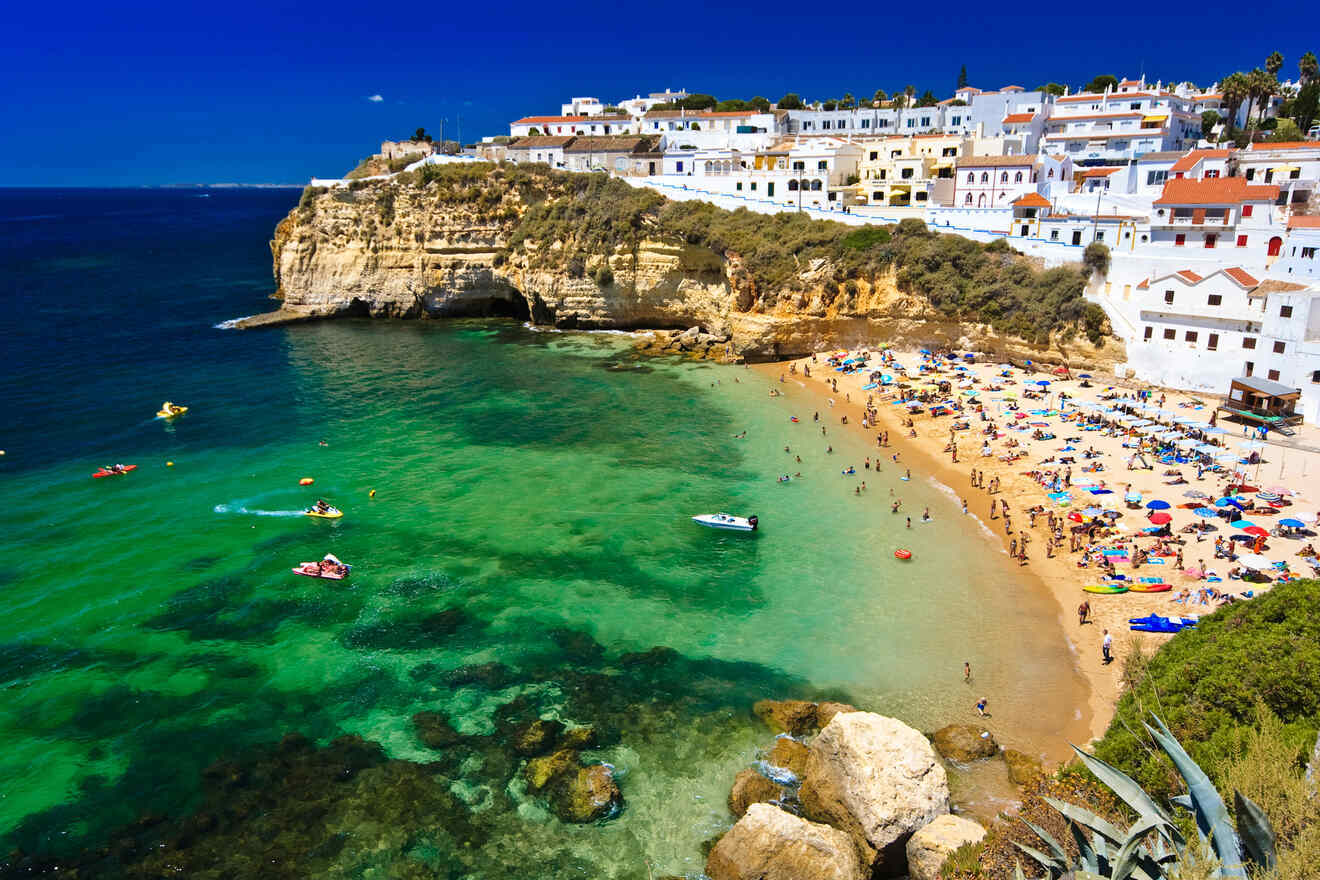 Bustling beach scene in Algarve with clear turquoise waters, flanked by rugged cliffs and white Mediterranean-style buildings under a bright blue sky.