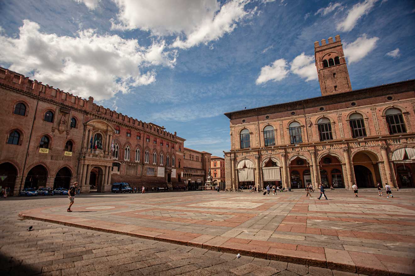 Spacious Piazza Maggiore in Bologna under a blue sky with clouds, showcasing the grandeur of surrounding medieval and renaissance architecture.
