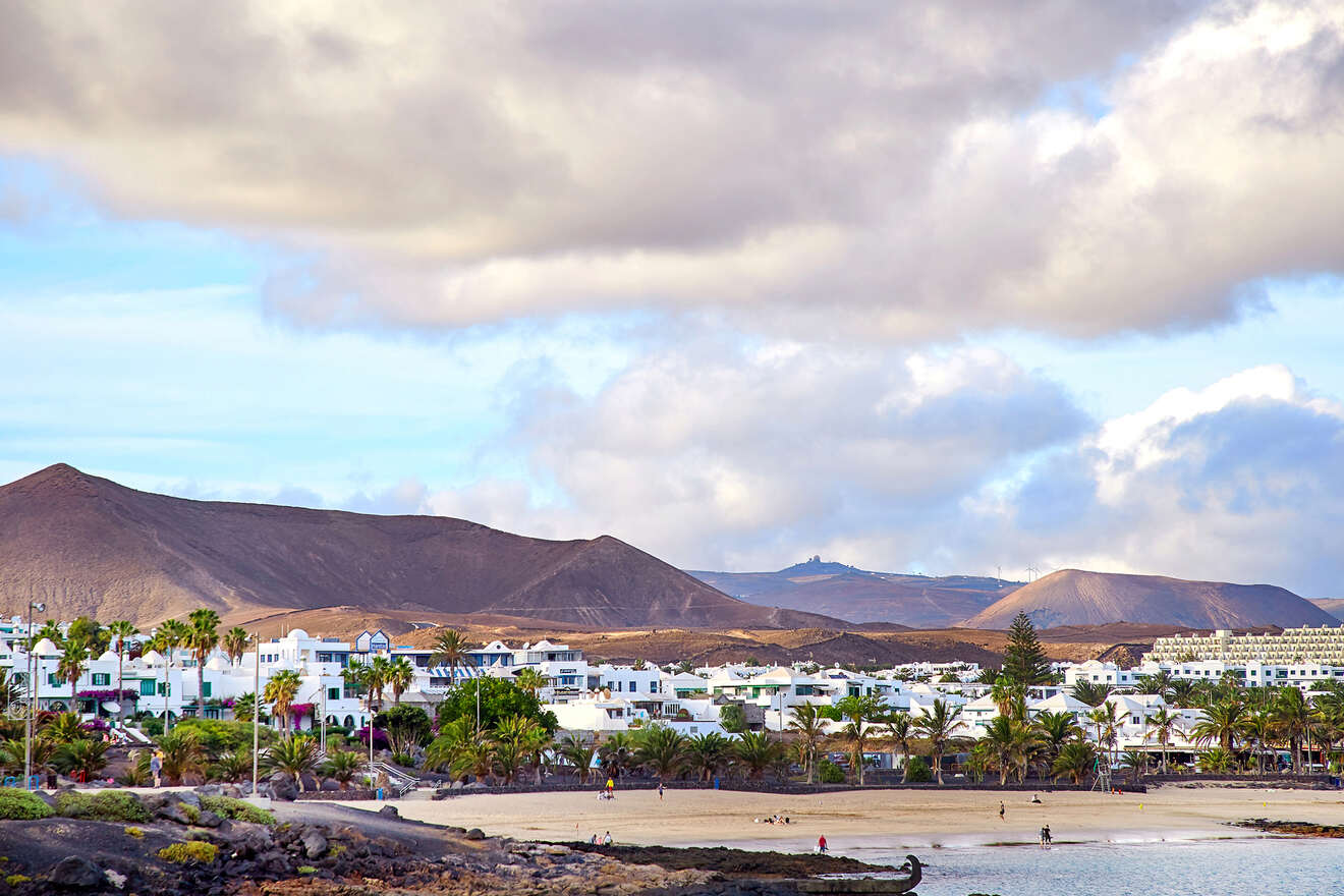 Serene beachfront scene in Lanzarote with palm trees, white houses against the backdrop of volcanic hills, under a cloudy sky.