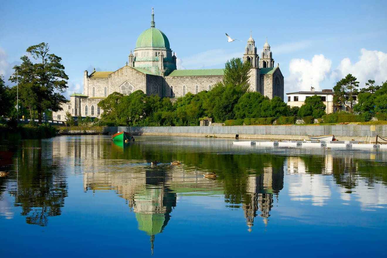 Historic stone cathedral with a green dome, reflected in the still water of a river, with a backdrop of trees and clear skies.