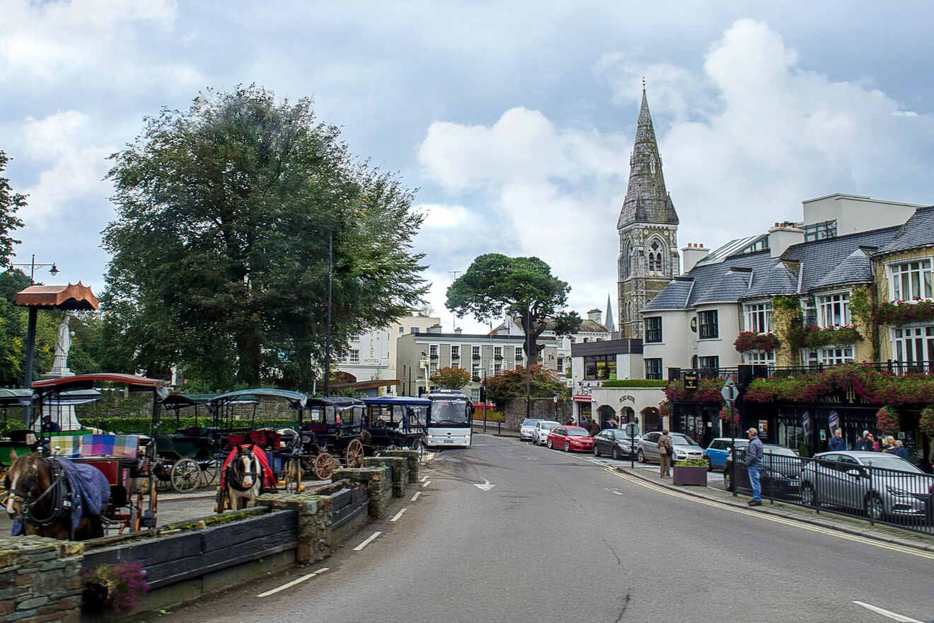 Historic street scene in Killarney with horse-drawn carriages, a church spire, and flowering balconies on a cloudy day