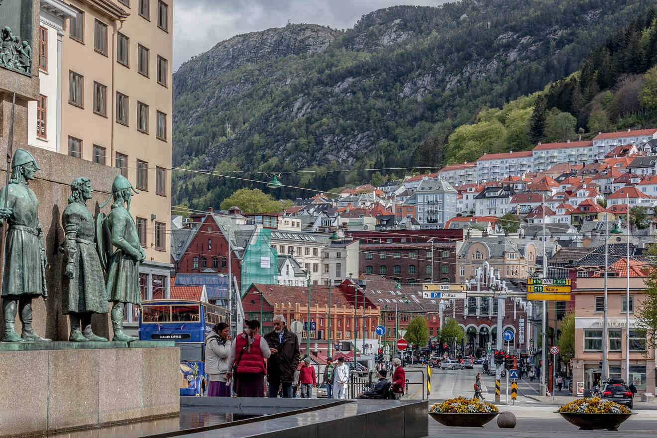 A bustling city scene in Bergen, Norway, showcasing the dynamic contrast between historical statues in the foreground, lively urban streets filled with people and vehicles, and the serene backdrop of forested mountains