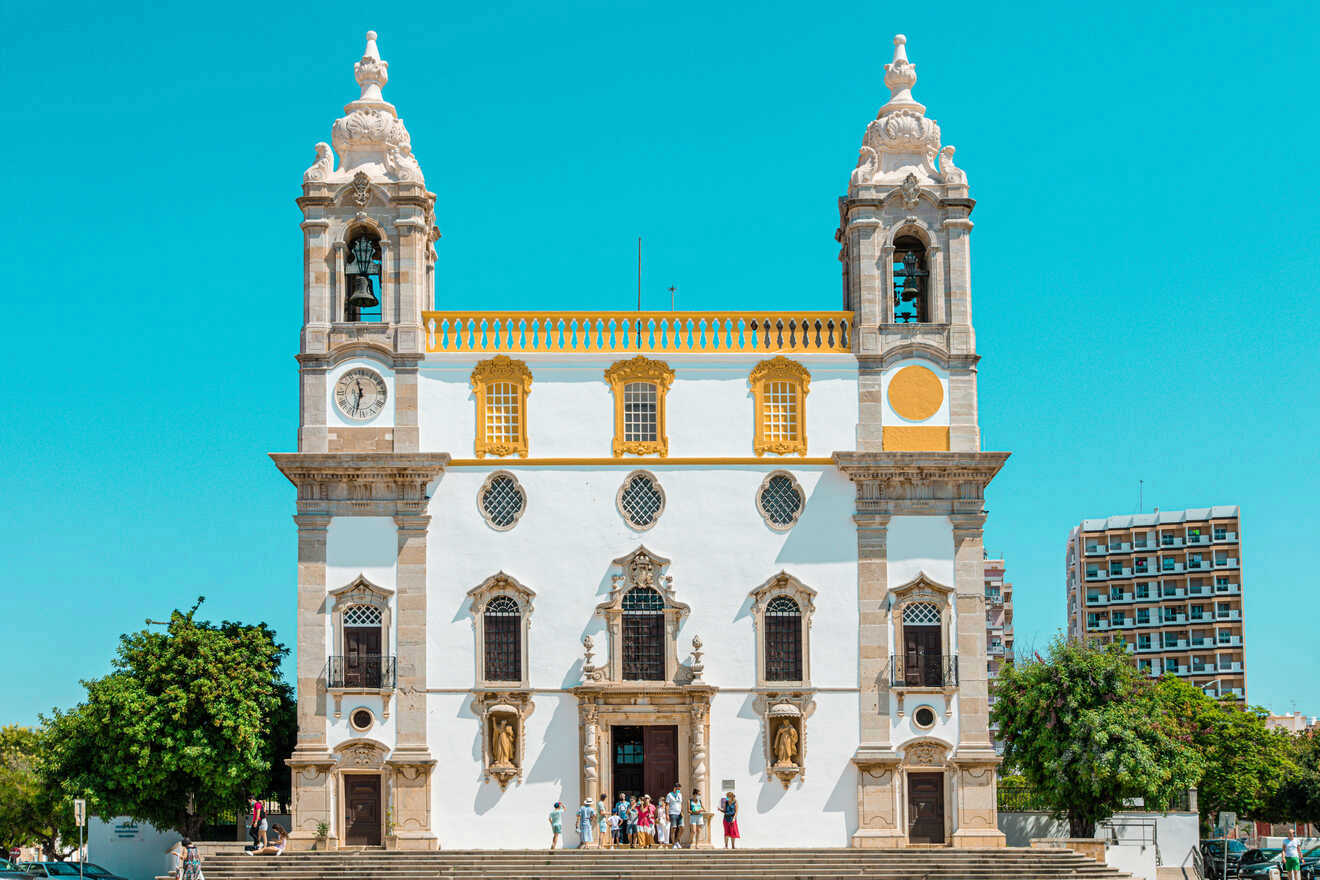 Historic church facade with ornate Baroque architecture, twin bell towers, and visitors at the entrance, set against a clear blue sky