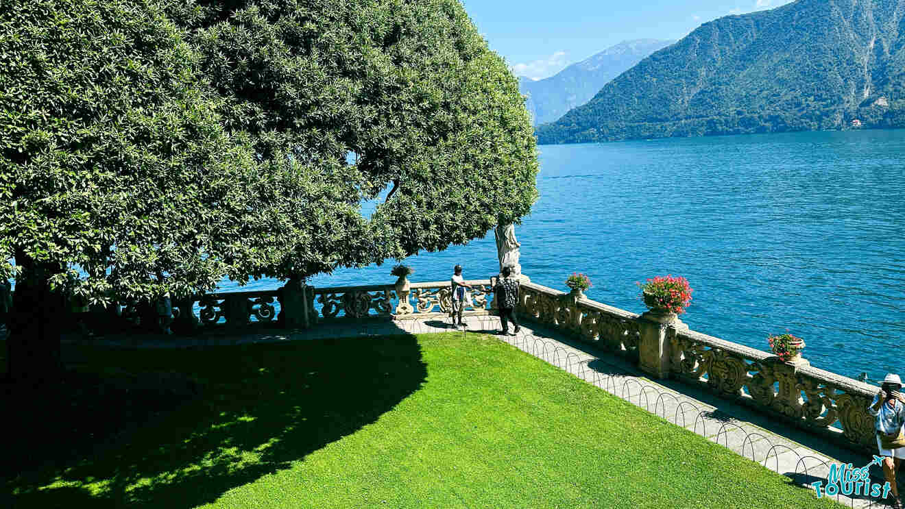 Ornate lakeside garden with manicured trees and a hedge-lined pathway, overlooking the serene blue waters of Lake Como