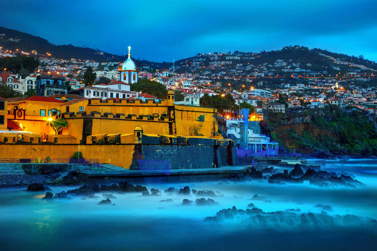 A coastal townscape at twilight with the glowing lights of buildings, a church spire, and the long exposure smoothing the sea into a misty surface