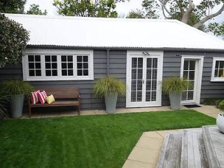 A quaint grey guesthouse with white trim, nestled in a well-kept garden with a bench and potted plants