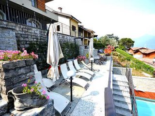 Stone terrace by a pool with white lounge chairs, umbrellas, and blooming flowers, offering a relaxing space with a view of Lake Como hills.