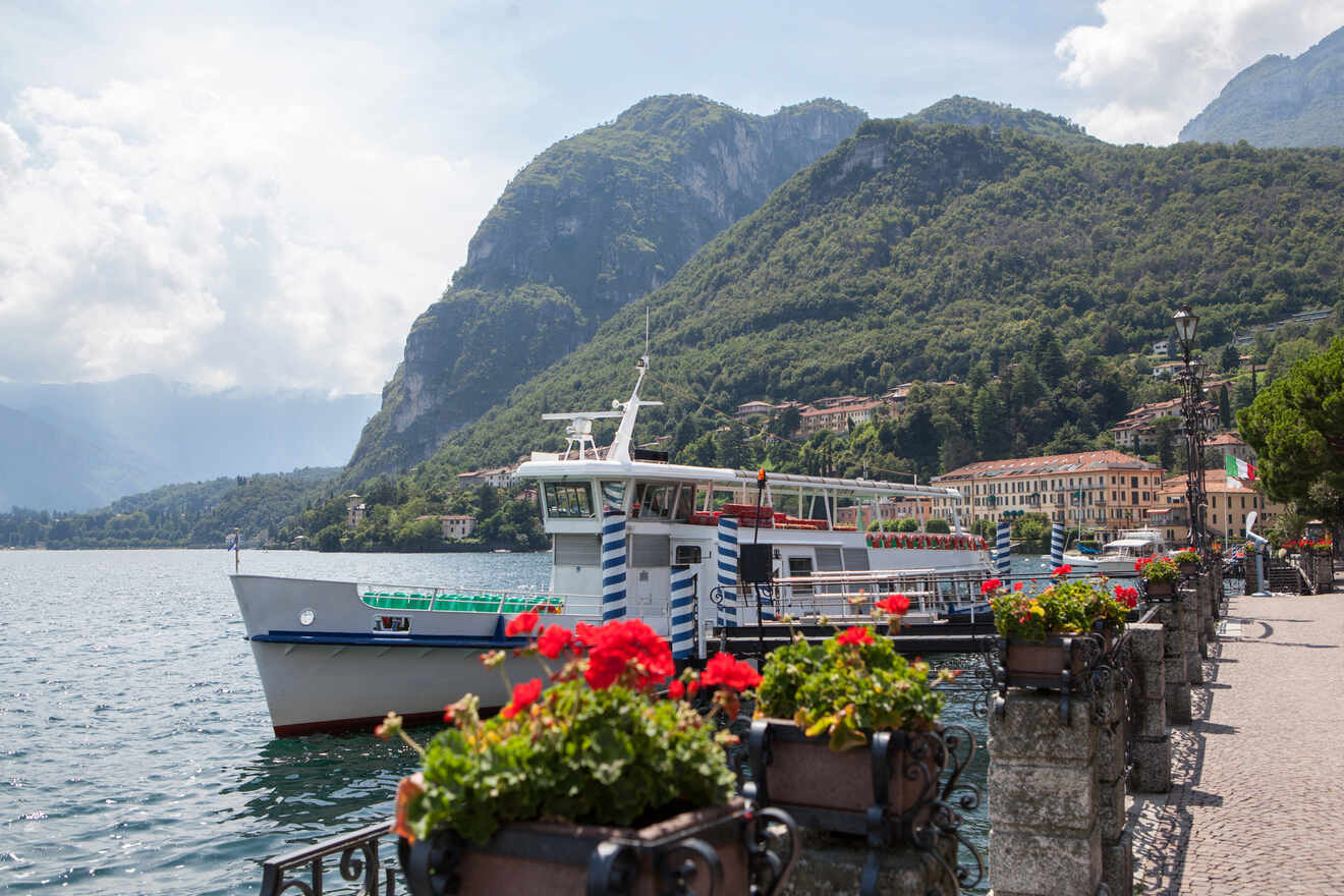 A picturesque lakeside promenade in Lake Como, with a moored ferry boat, vibrant flower beds, and a backdrop of mountains and Italian villas