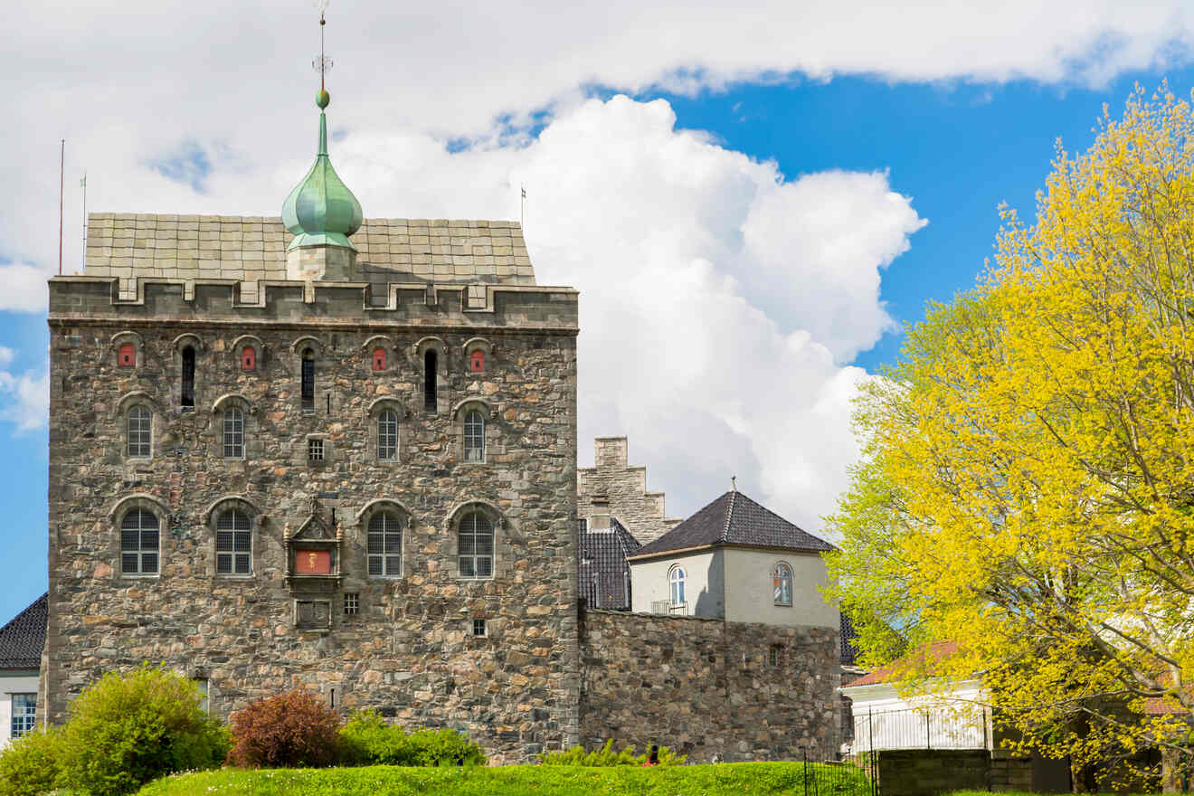 The historic Rosenkrantz Tower in Bergen, Norway, stands against a bright sky, its stone facade and distinctive green turret surrounded by vibrant spring foliage