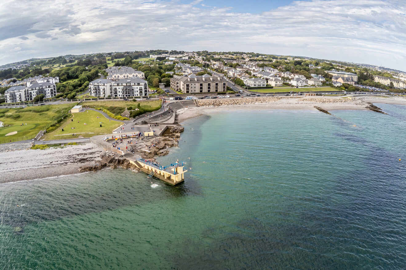 Aerial view of a coastal town with apartment buildings, a beach, and a pier, with clear blue waters and a green landscape surrounding it.