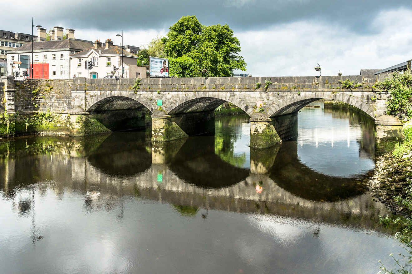 An old stone bridge in Cork, Ireland reflecting in the water with surrounding greenery and urban backdrop.