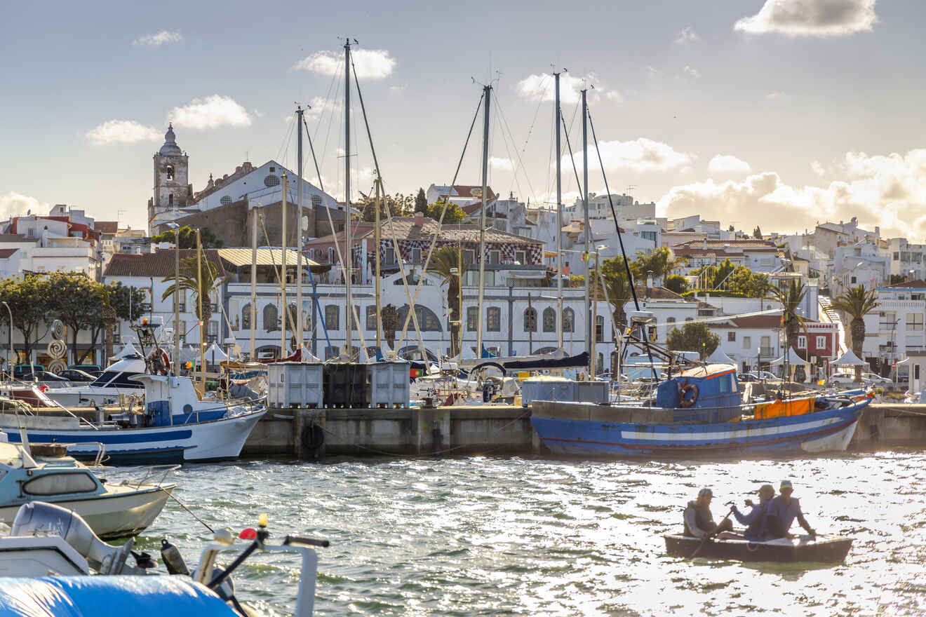 Quaint marina scene with boats moored in the harbor, a group of people rowing in the foreground, and the charming white architecture of a coastal town under a soft-glowing sun