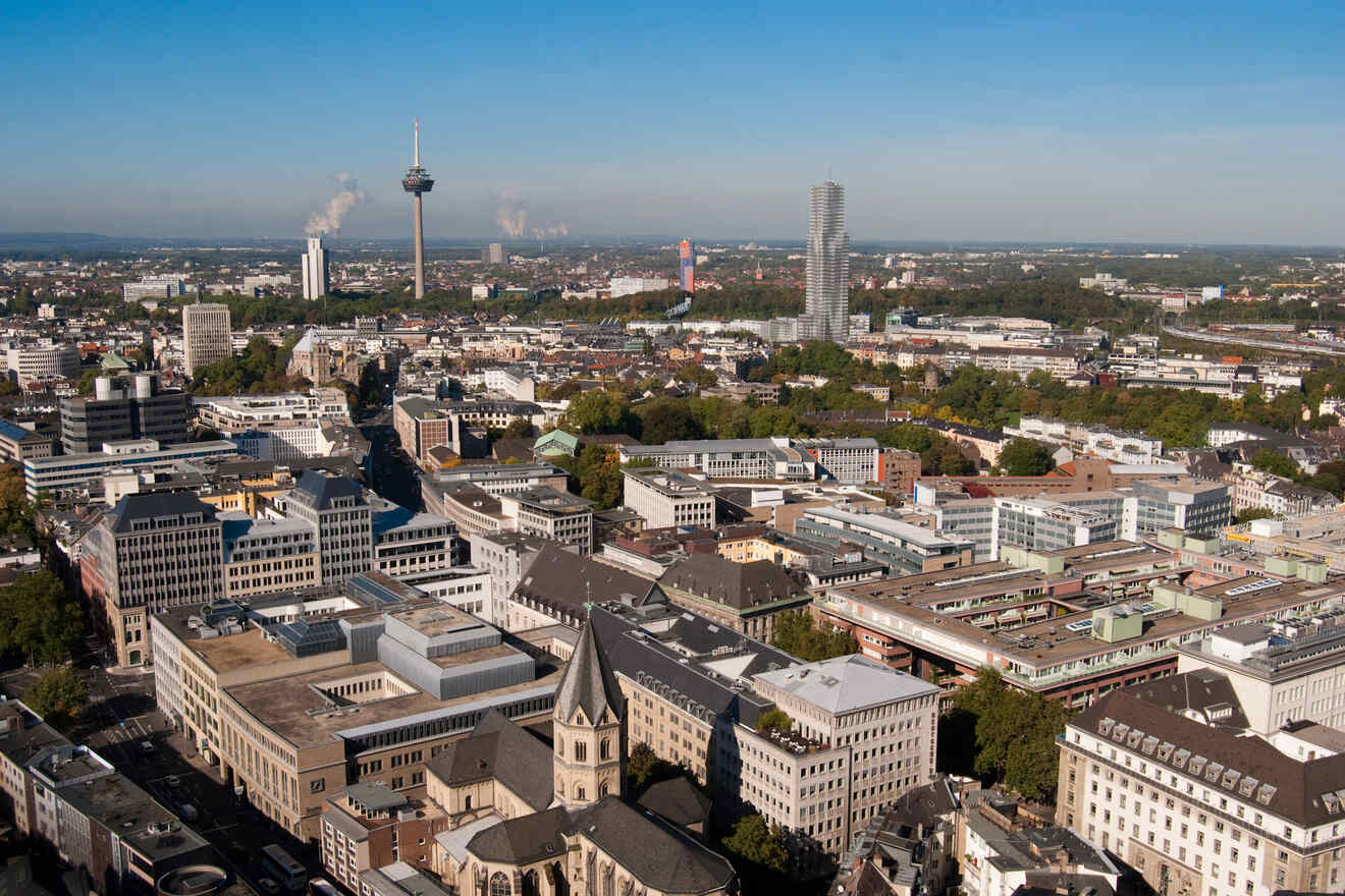 A cityscape of Cologne featuring the Colonius Tower, KölnTriangle, and a sprawling urban layout with various buildings and the central railway lines.