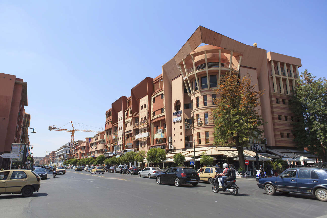 Busy street scene in Marrakech with cars and motorcycles in front of modern terracotta-colored buildings under a clear blue sky