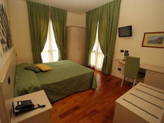 Simplistic hotel room with a double bed covered in green patterned bedding, light wooden floors, and basic amenities against a soft cream wall.