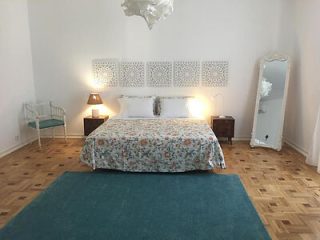 Minimalist bedroom with a queen-sized bed, white walls, patterned bedspread, and a teal accent rug on a herringbone floor
