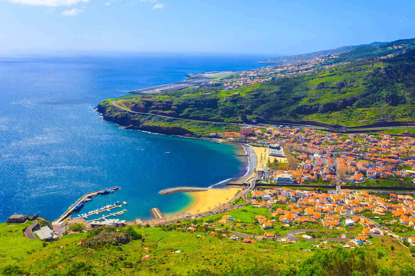 A bird's-eye view of a coastal town with a crescent-shaped beach, a marina, and a runway, surrounded by lush green terrain