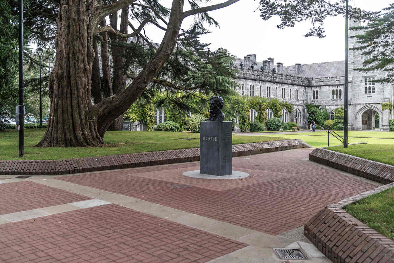 A statue of Boole in front of a university's lush green courtyard with gothic-style buildings in the background.