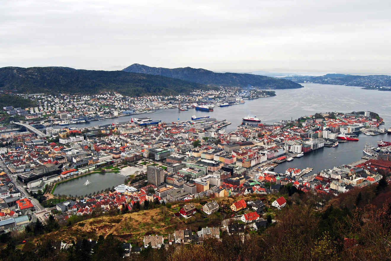 Panoramic view of Bergen, Norway, captured from a high vantage point, showcasing the sprawling cityscape, the extensive harbor with ships docked, and the surrounding mountains under a cloudy sky
