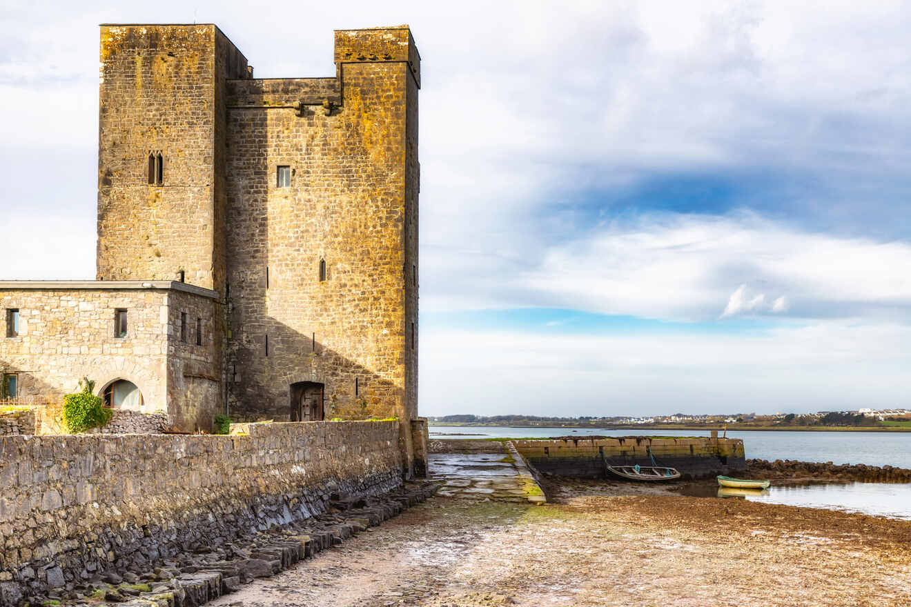 Medieval stone tower of a castle with a small wooden door, beside a dry moat with a boat, under a partly cloudy sky.