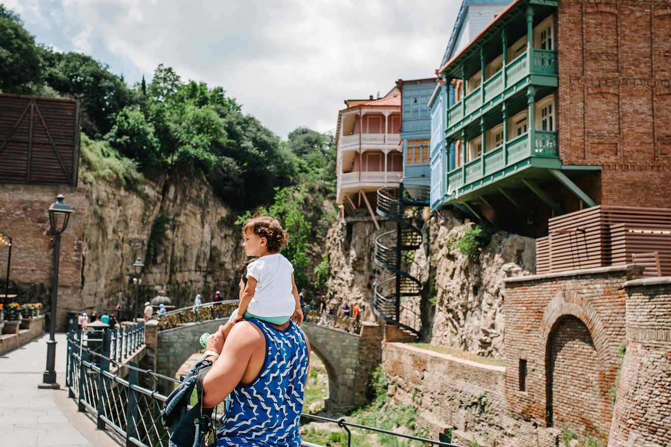 A mother carrying her child on her shoulders, with Tbilisi’s famous cliff-hanging houses in the background, reflecting a moment of family enjoyment in a historic setting