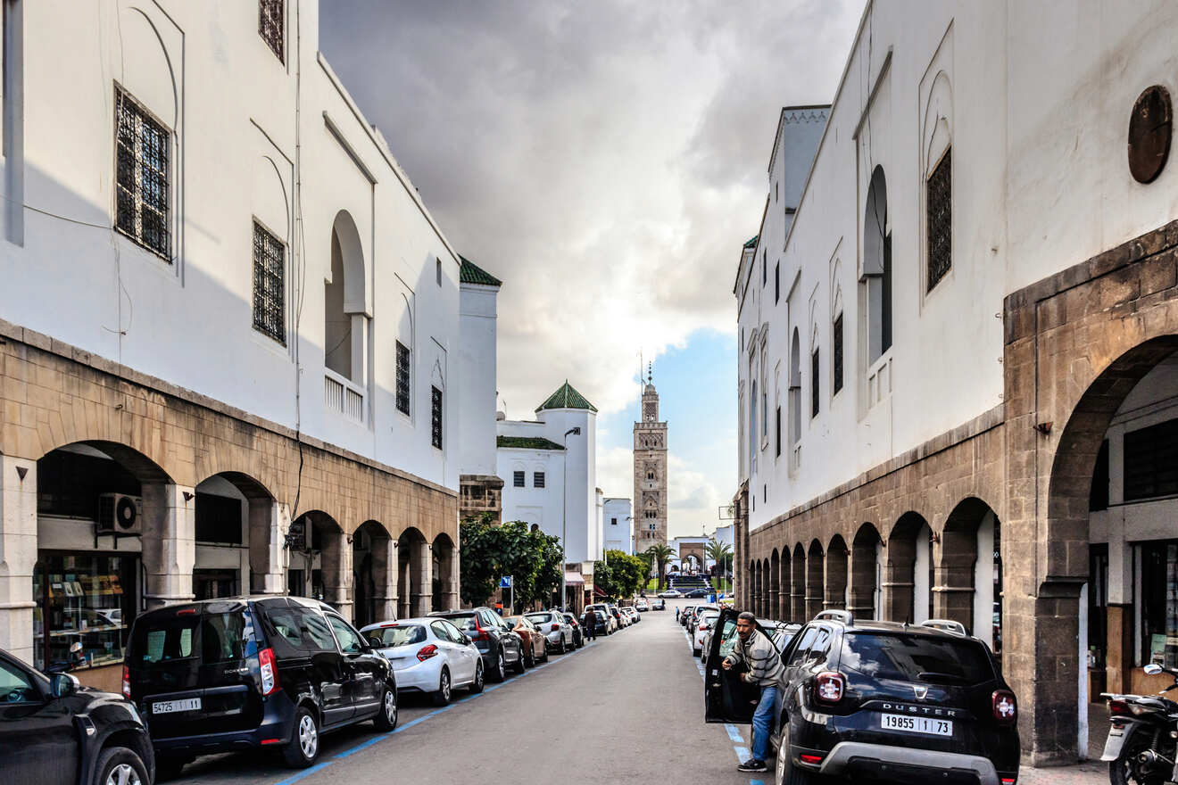 Historical Moroccan street with cars parked along the sidewalk, white buildings with arched details, and a distant mosque tower.